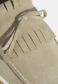 Lichtbeige suede schoen met een moccasin ontwerp. Heeft een franjerand en een dunne strik, en toont een gestructureerd oppervlak en contrasterende stiksels.