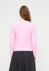 Πουλόβερ με μακριά μανίκια σε ανοιχτό ροζ χρώμα, κατασκευασμένο από απαλό ύφασμα, με στρογγυλή λαιμόκοψη και εφαρμοστή γραμμή, συνδυασμένο με σκουρόχρωμη πλισέ φούστα.