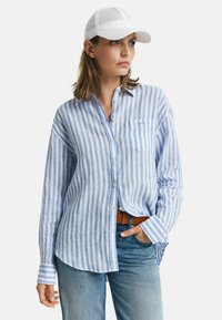 Camicia a righe azzurro chiaro e bianca con una tasca, abbinata a jeans in denim chiaro e un cappellino bianco.