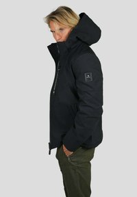 Veste isolante noire avec un col montant et une capuche, dotée d'une fermeture zippée, de poches latérales et d'un patch logo sur la manche. Texture lisse.