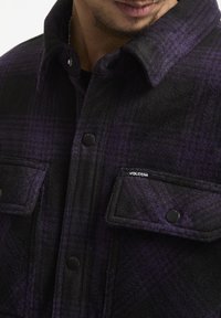 Chaqueta de franela de cuadro negro y púrpura con cierre de botones, dos bolsillos en el pecho y una etiqueta de Volcom en el bolsillo izquierdo. Textura suave.