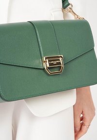 Borsa a tracolla in pelle verde con texture, dotata di una chiusura in hardware dorato, caratterizzata da un design a pattina triangolare e una tracolla regolabile per un utilizzo versatile.