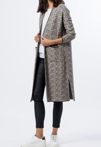 Długi, wzorzysty płaszcz w neutralnych odcieniach z bocznymi rozcięciami. Charakteryzuje się prostą sylwetką, otwartym przodem i dwoma przednimi kieszeniami. Połączony z czarnymi spodniami i białymi trampkami.