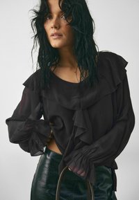 Blusa nera arricciata con maniche lunghe e polsini arruffati, abbinata a una gonna in pelle verde lucida e a una borsa piccola.