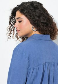 Camicia azzurra con texture, colletto e pieghe posteriori, dotata di maniche lunghe. Accompagnata da grandi orecchini a cerchio metallici.