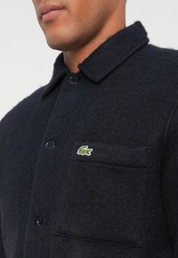 Zwarte overhemdjas van een wolmix met een puntkraag, knopen aan de voorkant, een borstzak en een groen geborduurd krokodillenlogo.