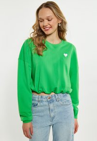 Zielony, krótki sweatshirt z długimi rękawami, z małym białym sercem na piersi. W połączeniu z jasnoniebieskimi spodniami o wysokim stanie.