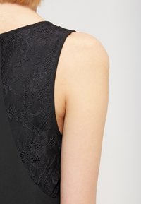 Černý top bez rukávů s texturovanou krajkovou zadní částí, hladkým materiálem vpředu a zaobleným výstřihem na ramenním švu.