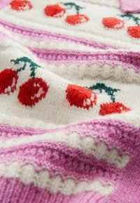 Gebreide deken met een witte basis, rode kersenmotieven, roze strepen en textuurrijke patronen, die samen een gezellige en levendige uitstraling geven.