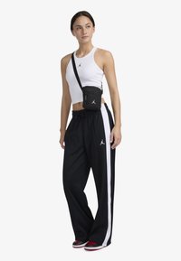 Top scurt alb, pantaloni negri de trening cu dungi albe pe lateral și o geantă mică neagră purtată în diagonală, toate având logo-ul Jordan. Pantofi roșii.