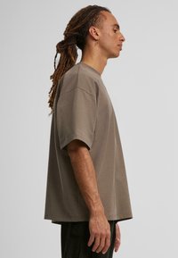 Př oversized tmavě hnědém bavlněném tričku s krátkým rukávem, které má klasický kulatý výstřih a volný střih, zobrazeném ze strany.