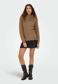 Pull marron à col bénitier en tissu tricot doux, avec un ourlet côtelé, porté sur une jupe à motif sombre et associé à des bottes noires hauteur genou.