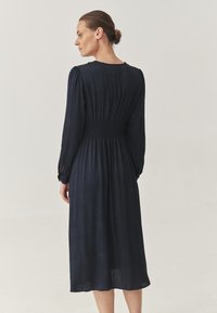 Vestido azul marino de manga larga con cintura fruncida, de longitud media, tejido texturizado y patrones sutiles. Presenta detalles de puños abotonados.