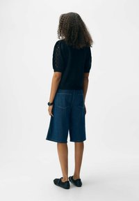Personne avec des cheveux bouclés portant un haut noir à manches courtes et un short en denim jusqu'aux genoux, se tenant de dos sur un fond blanc.