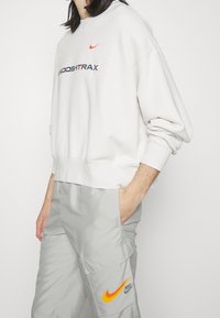 Bílá mikina s kulatým výstřihem a dlouhými rukávy, na přední straně s barevným logem a textem. Spárována se světle šedými cargo kalhotami.