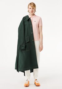 Grön trenchcoat med slagknappar draperad över en blek rosa polo. Vita byxor med elastiska manschetter och orange sneakers.
