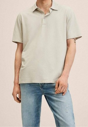 Mężczyzna ubrany w jasnobeżową koszulkę polo z krótkim rękawem i niebieskie dżinsy, stojący w luźnej postawie, jasna karnacja, tatuaż na lewej ręce.