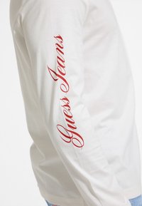 White long-sleeve shirt made of cotton. Features "Guess Jeans" printed in red cursive down the left sleeve. Simple, smooth texture.