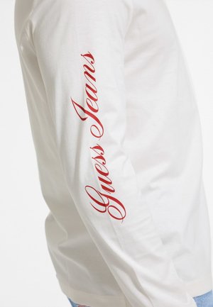 Balta ilga rankovė marškiniai, pagaminti iš medvilnės. Kairėje rankovėje raudonos kursyvo raidėmis parašyta "Guess Jeans". Paprasta, lygi tekstūra.