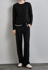 Osoba ubrana w czarny sweter z białą lamówką, czarne spodnie z jedną ręką w kieszeni oraz białe trampki, stojąca na tle szarej ściany.