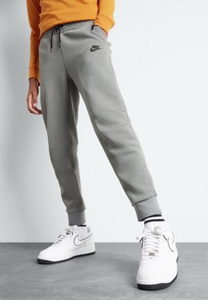 Osoba ubrana w szare, teksturowane spodnie dresowe Nike, białe buty sportowe Nike z czarnym logo, czarno-białe paskowane skarpetki oraz pomarańczową bluzę.
