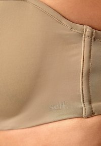Beige bh met ribbeltextuur, naadloos ontwerp en zichtbaar merkembleem. Materiaal lijkt zacht en rekbaar, met haak-en-oogsluiting.