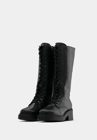 Ζεύγος μαύρων δερμάτινων μποτών μέχρι το γόνατο με κορδόνια και χοντρό τακούνι σε λευκό φόντο.