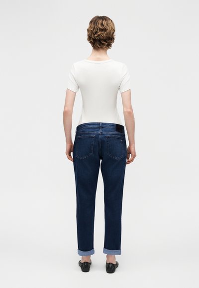 Άτομο με κοντά σγουρά μαλλιά στέκεται με την πλάτη γυρισμένη, φορώντας λευκό κοντομάνικο πουκάμισο, σκούρα μπλε τζιν με ρεβέρ και μαύρες μπαλαρίνες.