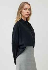 Blouse en soie noire avec un col et une coupe ample, associée à une jupe à carreaux gris clair présentant des lignes et textures discrètes.