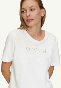 Weißes Baumwoll-T-Shirt mit kurzen Ärmeln, Rundhalsausschnitt und einem strukturierten "VOYAGE"-Druck auf der Brust. Glatter Stoff, lässige Passform.