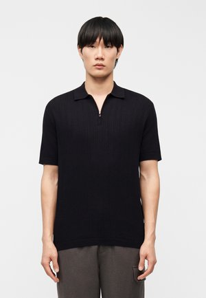 Mann mit glatten schwarzen Haaren trägt ein schwarzes, kurzärmeliges Strick-Poloshirt mit einem Reißverschluss vorne und graue Hosen, steht vor einem einfarbigen Hintergrund.