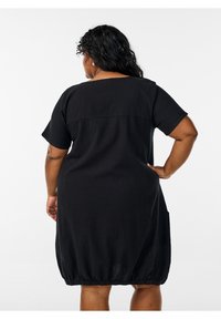 Sieviete ar cirtainiem melniem matiem, tērpusies īsu piedurkņu melnā kleitā ar elastīgu apmali, stāv ar vienu roku uz gūžas, skatoties prom.