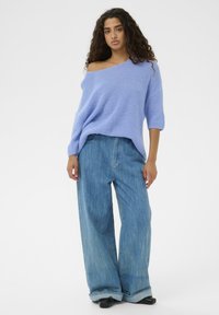 Pull en maille bleu clair surdimensionné avec un design épaules dénudées, associé à un jean denim bleu délavé ample et des chaussures noires.