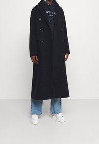 Cappotto doppiopetto in misto lana nera con ampi rever, due tasche frontali, indossato sopra un hoodie blu navy e jeans svasati blu medio con sneakers bianche.