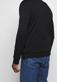 Sudadera negra de material suave, con cuello redondo y puños acanalados. Combina con pantalones vaqueros azules, mostrando un ajuste relajado.