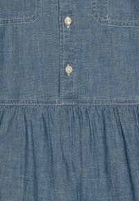 Džínové šaty s knoflíkovou légou vpředu, světle modré barvy, se dvěma náprsními kapsami a nařaseným pasem. Měkká textura a neformální design.
