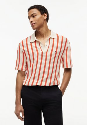 Mladý muž v béžovém polokošili s vertikálními oranžovými pruhy a černými kalhotami, stojící s jednou rukou v kapsě a dívající se stranou.