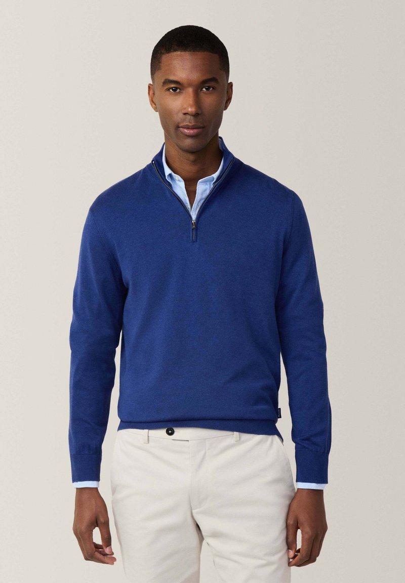 Pullover a collo zip blu realizzato in morbida lana, con maniche lunghe e orlo a costine, indossato sopra una camicia azzurra con colletto.