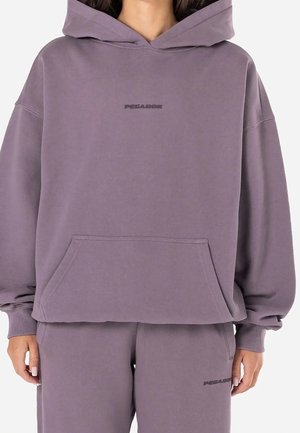 Frau trägt einen locker sitzenden lila Hoodie und passende Sweatpants mit einem kleinen "PEGADOR"-Logo auf Brust und Oberschenkel.