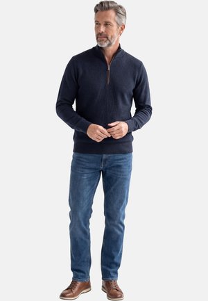 Man met grijs haar en baard, gekleed in een marineblauwe trui met een kwart-ritssluiting, blauwe spijkerbroek en bruine leren schoenen, staand tegen een effen achtergrond.