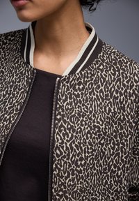 Persona che indossa una maglietta nera e una giacca con stampa leopardata e cerniera, con colletto a righe bianche e nere, su sfondo semplice.