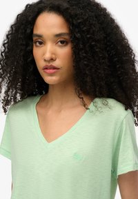 Camiseta de cuello en V verde claro hecha de una tela texturizada, con mangas cortas y un pequeño logo bordado en el pecho.