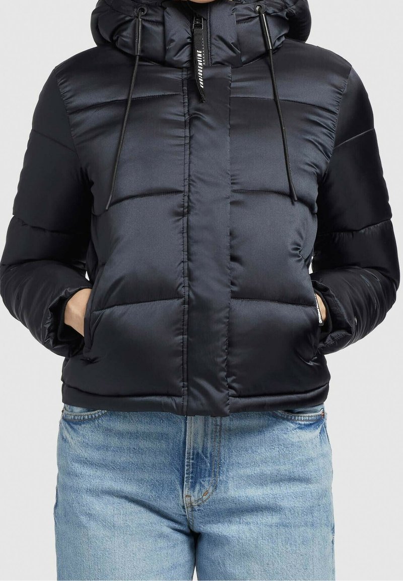 Veste noire matelassée avec capuche et fermeture éclair frontale, mains dans les poches, associée à un jean denim bleu clair.
