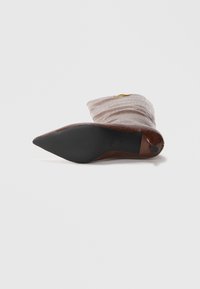 Chaussure brune à bout pointu fabriquée en matériau texturé, ornée d'un motif géométrique lisse et d'un petit accent doré sur le côté. Semelle noire.