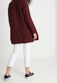 Burgundy stickad cardigan med lös passform, ribbat fåll och långa ärmar, kombinerad med vita cropped jeans och svarta sneakers.