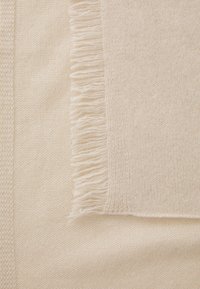 Couverture de couleur crème avec une texture douce. Avec des bords à franges et un motif tissé sur les côtés, offrant une sensation lisse et moelleuse.
