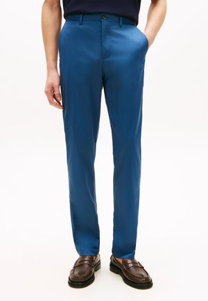 Man wearing blue tailored trousers with front pockets and dark brown leather loafers, standing against a plain white background.