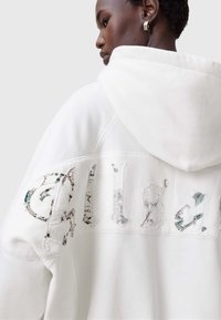 Sudadera blanca con capucha que presenta bordados texturizados en la parte posterior con detalles de técnicas mixtas. Tela suave, ajuste relajado y capucha ajustable.