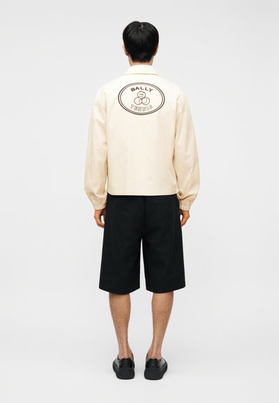 Άνδρας με εκρού μπουφάν με το λογότυπο "Bally Tennis" στην πλάτη, μαύρο σορτς μέχρι τα γόνατα και μαύρα αθλητικά παπούτσια, όρθιος με την πλάτη προς τα εμπρός.