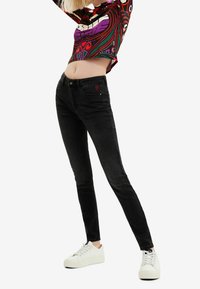 Černé skinny džíny s těsným střihem, se červeným srdíčkovým logem na kapse. Kombinované s barevným vzorovaným zkráceným topem a bílými teniskami.